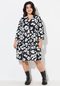Schwarz-weißes A-Linien-Kleid mit knopflosem Verschluss, 3/4-Ärmeln, Blumenmuster und gerafftem Taillenbereich, kombiniert mit schwarzen, mit Nieten besetzten Stiefeletten.
