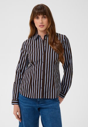 Femme aux longs cheveux bruns portant une chemise noire à boutons avec des rayures verticales blanches et bleues et un jean bleu, debout devant un fond blanc.