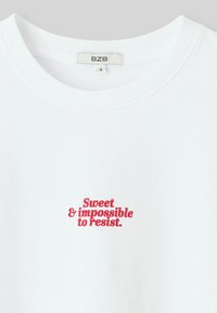 T-shirt en coton blanc avec un col rond, comportant un texte rouge "Doux et impossible à résister" à l'avant. L'étiquette de taille est visible.