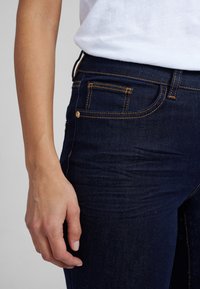 Jeans de mezclilla azul oscuro con costuras naranjas y un detalle en el bolsillo delantero. La tela parece ligeramente texturizada, ajustándose al cuerpo.