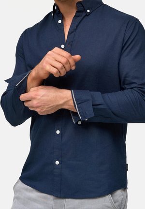Homme ajustant les poignets d'une chemise bleu marine à manches longues boutonnée, portant un pantalon gris clair, fond uni.