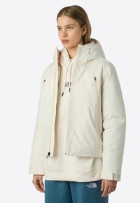 Ung person som bär en off-white huva pufferjacka �över en krämfärgad hoodie och tealfärgade sweatpants, står mot en enkel bakgrund.