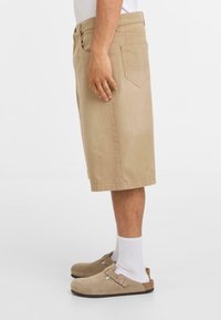 Beige katoenen shorts met een rechte snit, tot op de knie, met een achterzak en een metalen knoopsluiting. Gecombineerd met lichtgekleurde klompen en sokken.