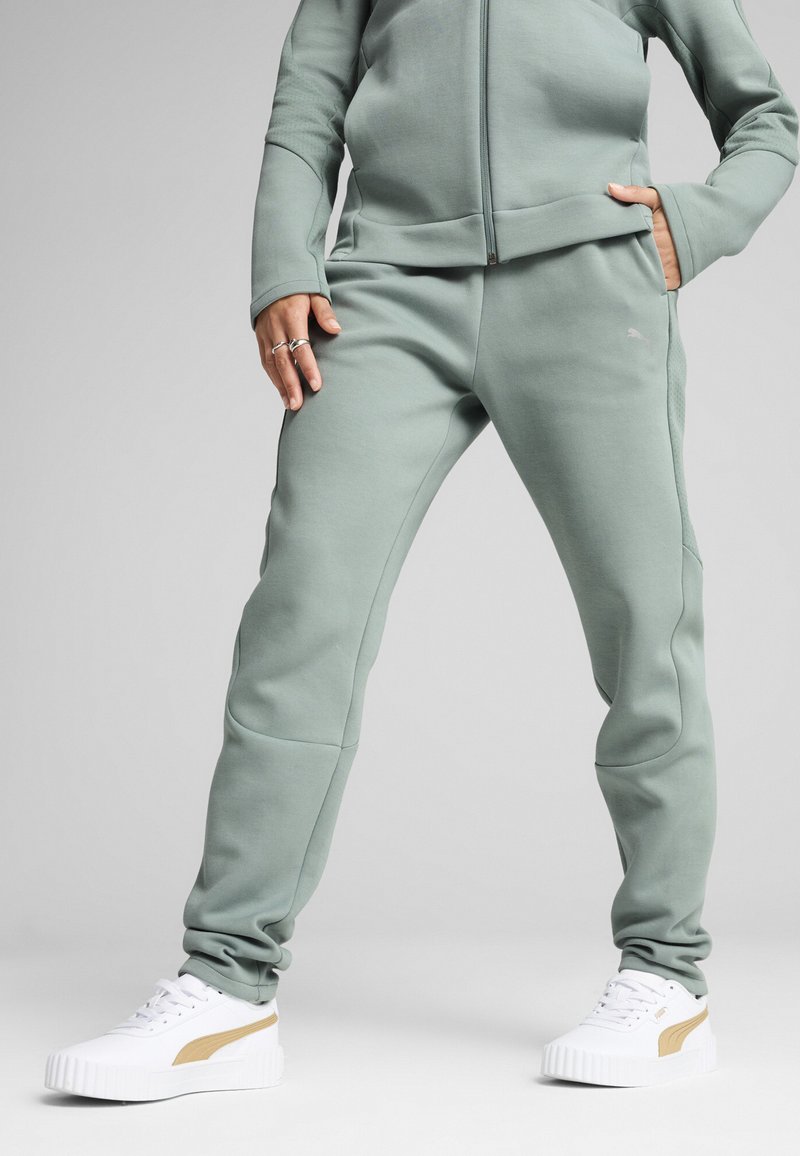 Ensemble de jogging vert clair comprenant une veste zippée et un pantalon fuselé avec des accents texturés, associé à des baskets blanches portant un logo doré.