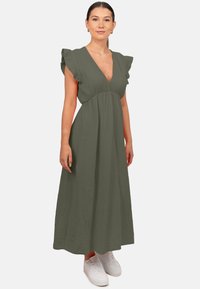 Robe maxi vert olive avec un décolleté en V profond, des manches volantes et un tissu texturé. Portée avec des baskets blanches.