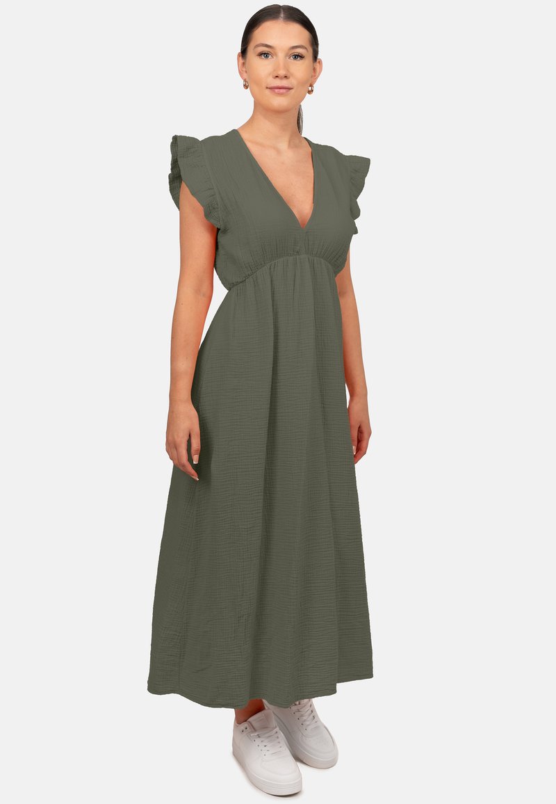 Robe maxi vert olive avec un décolleté en V profond, des manches volantes et un tissu texturé. Portée avec des baskets blanches.