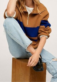 Pull en rayures marron et bleu avec un col zippé court, porté avec un jean léger bleu délavé et des mocassins noirs, reposant sur un bloc en bois.