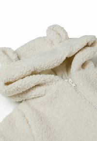 Veste en polaire couleur crème avec un extérieur texturé et doux. Dispose d'une capuche avec des oreilles et d'une fermeture éclair sur le devant.