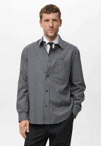 Camisa de cuadros gris con cuello puntiagudo, que cuenta con un bolsillo y mangas largas. Llevada sobre una camisa blanca y una corbata.