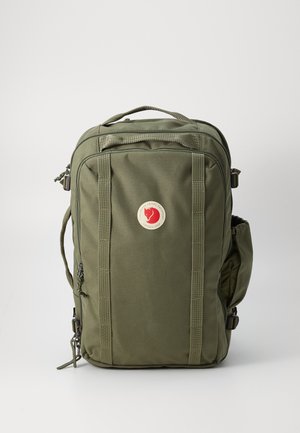 Sac à dos vert olive avec un patch logo, plusieurs zips, détails sur le panneau avant et poche latérale, fabriqué en tissu durable et avec un design structuré.