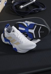 Scarpe da ginnastica bianche e blu con strisce beige posizionate su una superficie nera, con cavo nero arrotolato sullo sfondo.