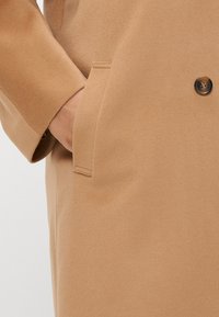 Καφέ παλτό από μείγμα μαλλιού με αυστηρό γιακά, κλείσιμο με ένα κουμπί και πλαϊνές τσέπες. Μαλακή υφή με λεία επιφάνεια.