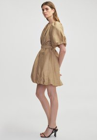 Tan linnen jurk met een diepe V-hals, pofmouwen en een riem om de taille. Glad textuur en een uitlopende rok, gecombineerd met zwarte enkelbandhakken.