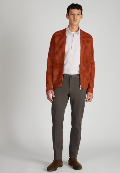 Cardigan arancione con texture sopra una camicia a righe chiara, abbinato a pantaloni scuri e stivali in suede marroni, su uno sfondo neutro.