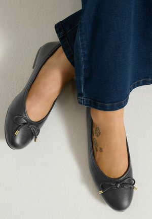 Pieds portant des chaussures de ballet basses en cuir gris avec détail nœud, associées à un pantalon en denim bleu foncé, tatouage visible à la cheville d'un pied.