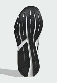 Čierna gumová podrážka obuvi s textúrovaným vzorom, obsahujúca predĺžené drážky a biele akcenty, navrhnutá pre trakciu a stabilitu.