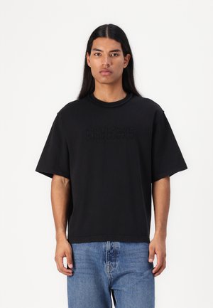 Must have lühikeste varrukatega must T-särk, millel on ümar kaelus ja esiküljel reljeefne "GUESS ORIGINALS" logo, koos helesiniste denim teksadega.