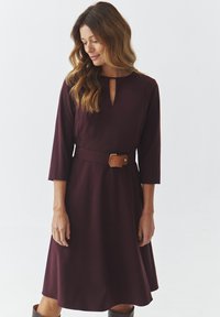 Robe A-line bordeaux avec un décolleté en trou de clé, des manches trois-quarts et une ceinture beige. Tissu doux avec une texture lisse.