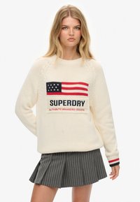 Crèmekleurige gebreide trui met een rode, witte en blauwe Amerikaanse vlag ontwerp en "SUPERDRY" tekst. Gestreepte accenten op de mouwen.