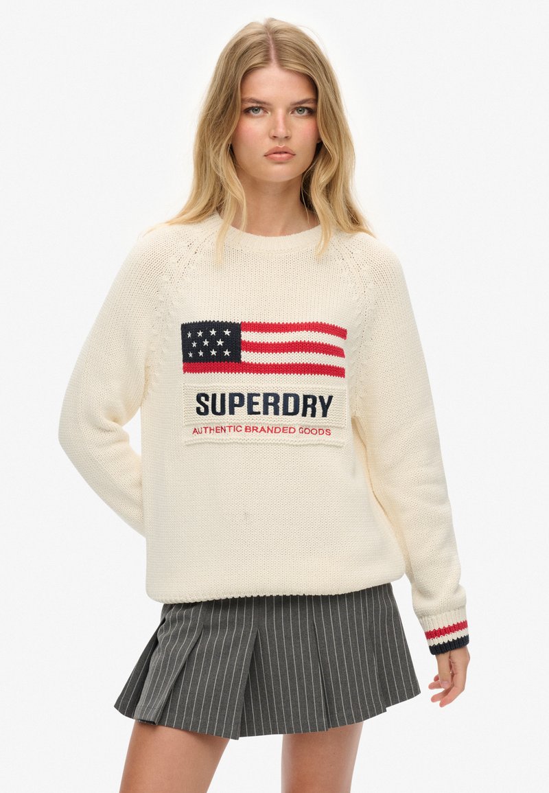 Crèmekleurige gebreide trui met een rode, witte en blauwe Amerikaanse vlag ontwerp en "SUPERDRY" tekst. Gestreepte accenten op de mouwen.