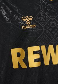Maillot de sport noir avec des motifs hexagonaux texturés. Texte "REW" en or et logo Hummel mis en avant en broderie dorée.