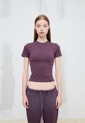 Mujer joven con el pelo largo y liso, con una camiseta morada ajustada y pantalones deportivos sueltos a juego, de pie contra una pared clara y lisa.