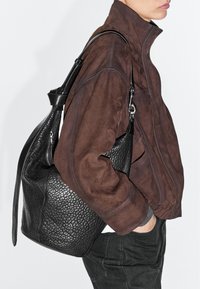 Personne en veste en daim marron portant un grand sac à main en cuir noir texturé sur l'épaule, avec la main dans la poche, sur fond blanc.