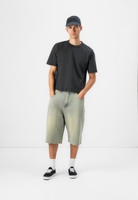 Tricou negru din bumbac, croială oversized, pantaloni scurți din denim de culoare verde deschis, până la genunchi, cu buzunare, asortați cu pantofi negri și o șapcă de baseball închisă.