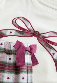 Vit topp med vinrött broderat bandmönster. Nedanför mönstrade byxor i vitt, grönt och vinrött med matchande rosettdetalj.