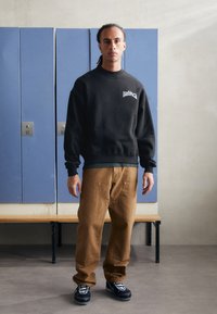 Black sweatshirt with logo, tan cargo pants, and black sneakers with white accents. Simple design and relaxed fit. Background features lockers.