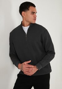 Tmavě šedý pulovr na čtvrtinový zip s vroubkovanou strukturou, který má dlouhé rukávy a stojáček. Model pózuje se zkříženýma rukama.