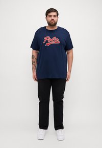 Jūras spēku kokvilnas T-kreklu ar izteiksmīgu sarkano "Polo" logo, brīvā griezumā, īsām piedurknēm, apvienots ar melniem biksēm un baltām sporta kurpēm.