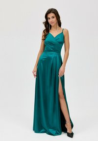 Tyrkysová saténová večerní toaleta s výstřihem do V, tenkými ramínky, nařaseným korpusem a vysokým rozparkem na stehně. Spárována s černými lodičkami se špičatou špičkou.