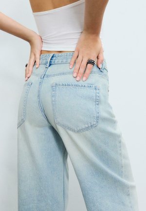 Oseba oblečena v svetlo modre široke kavbojke in beli kratki top, roke na boku, na eni roki črn prstan s srebrnimi zatiči.