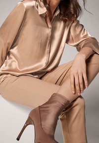 Blusa di satin beige con colletto, abbinata a pantaloni beige chiaro e stivali alla caviglia marroni con punta a punta e tacco alto.