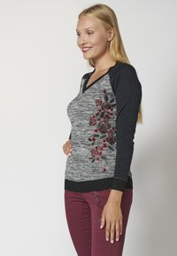Grauer und schwarzer Langarmpullover mit V-Ausschnitt, ausgestattet mit roten Blumenstickereien auf der linken Seite und einem gerippten Saum.