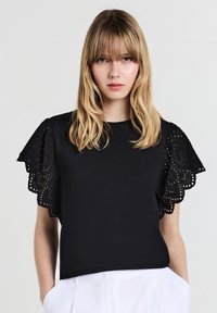 Giovane donna con capelli biondi indossa un top nero con maniche in pizzo e pantaloni bianchi, in piedi contro uno sfondo chiaro e semplice.