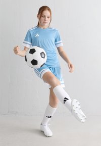 Junge Frau in hellblauem Fußballtrikot, die einen schwarz-weißen Fußball in einer Halle gegen eine weiße Wand tritt.
