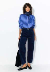 Chemise rayée bleue, pantalon en velours côtelé bleu marine, ballerines noires avec des clous, et un sac noir. Tissu lisse et coupe ample avec une ceinture accentuée.