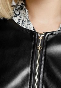 Chaqueta de cuero sintético negra con cremallera central y herrajes dorados, y un collar estampado debajo. Textura suave y diseño ajustado.