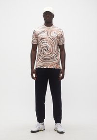 Άνδρας που φοράει μπλουζάκι με μπεζ και καφέ στροβιλώδες μοτίβο, μαύρο παντελόνι, λευκό καπέλο και λευκά αθλητικά παπούτσια, στέκεται μπροστά σε απλό λευκό φόντο.