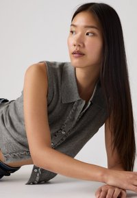 Νεαρή γυναίκα με μακριά σκούρα μαλλιά που φοράει αμάνικη γκρι πλεκτή μπλούζα, ξαπλωμένη στο πλάι, κοιτάζοντας προς τα αριστερά της σε απλό φόντο.