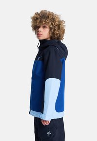 Niebieska i czarna kurtka z kapturem. Charakteryzuje się połączeniem materiałów i kolorów, w tym jasnoniebieskimi akcentami na rękawach. Logo na klatce piersiowej i kieszeni.