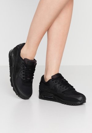 Nogi w czarnych butach Nike Air Max z siateczką i skórzanymi detalami, stojące na białej podłodze na tle szarego tła.