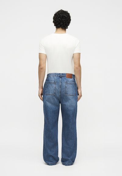 Άτομο με σγουρά μαύρα μαλλιά στέκεται πλάτη, φορώντας μια απλή λευκή κοντομάνικη μπλούζα και φαρδιά μπλε τζιν με πίσω τσέπες.