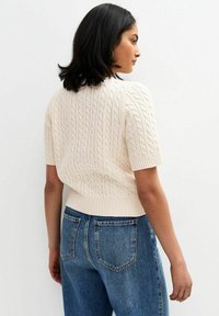 Krótki rękaw, kremowy sweter w warkocz z ribbanym wykończeniem, zestawiony z niebieskimi dżinsami o wysokim stanie z tylnymi kieszeniami. Teksturowane wykończenie na całej powierzchni.