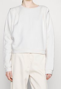 Person klädd i en enkel vit långärmad sweatshirt och vita högmidjade byxor, stående mot en neutral bakgrund.