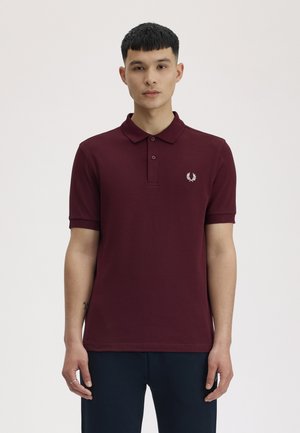Jeune homme portant un polo bordeaux à manches courtes avec un petit logo de laurier blanc sur la poitrine gauche, debout devant un fond uni.