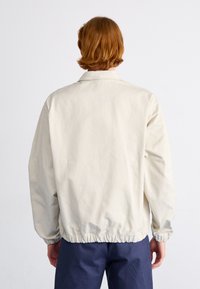 Lyse beige jakke med en afslappet pasform, der har en krave og elastisk kant. Materialet ser glat og let ud.