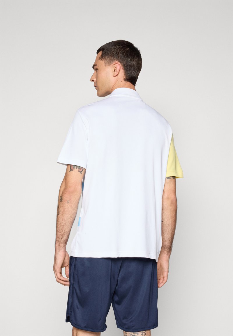 Polo à manches courtes de couleur blanche avec des accents jaunes et bleu clair, offrant une coupe décontractée et une texture lisse. Porté avec un short bleu marine.